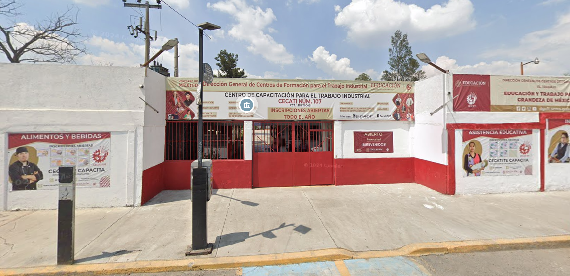 Fotografía en la que se muestra la entrada del plantel, conformada por muros que tienen los colores blanco y rojo. En medio de los muros hay un portón de color rojo que da acceso a las instalaciones. Arriba del portón hay una lona que contiene los datos del plantel, como el nombre y las carreras que se imparten.